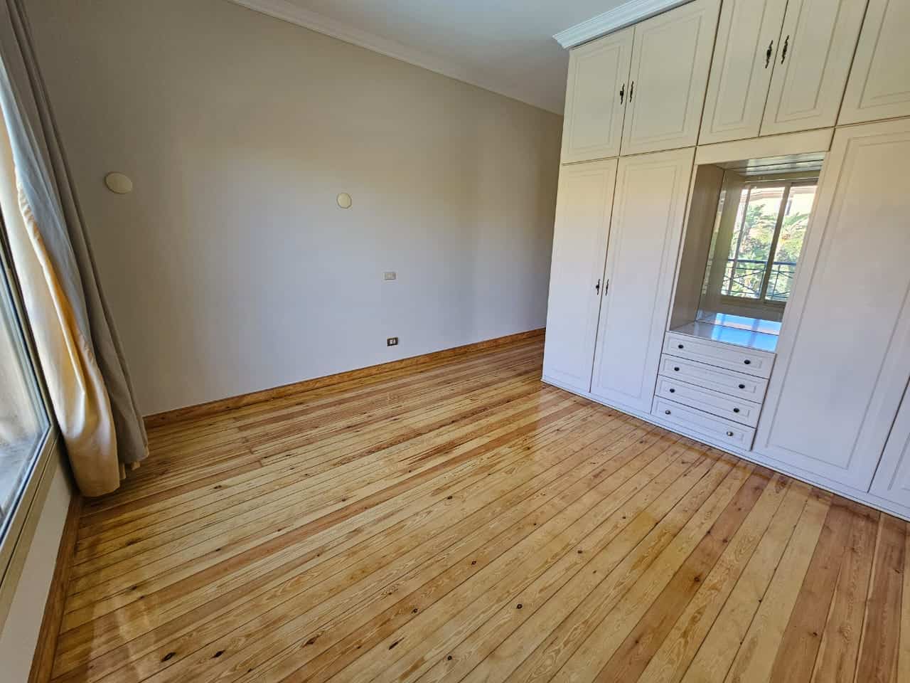 Bright empty bedroom with wooden flooring and built-in white wardrobe, large window with balcony view in Maadi Cairo.