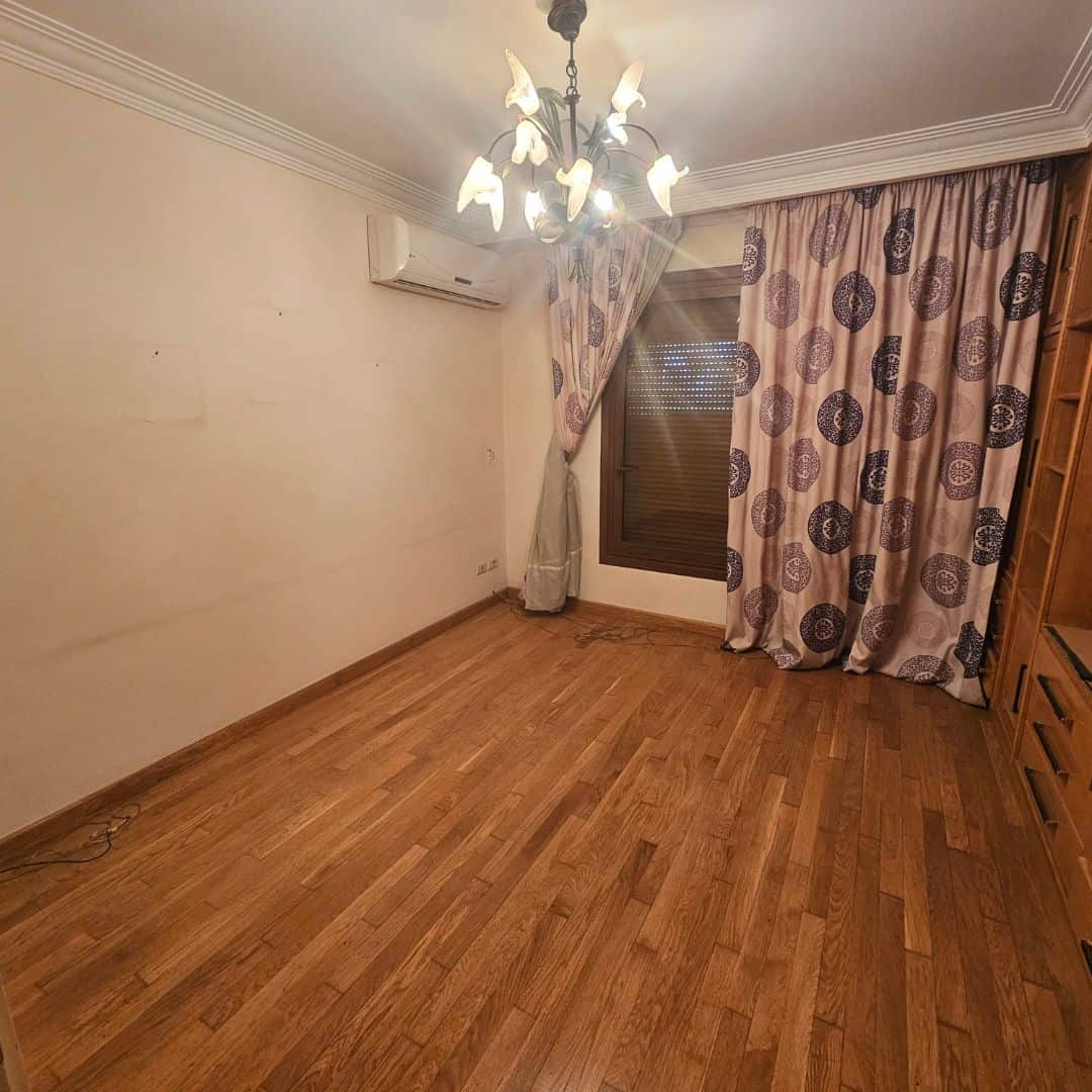 Spacious empty living room with wooden floors, chandelier lighting, and large window with patterned curtains.