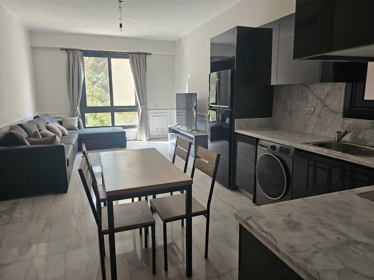 Modern apartment living room with large window, black sofa, and open kitchen in Maadi Cairo.