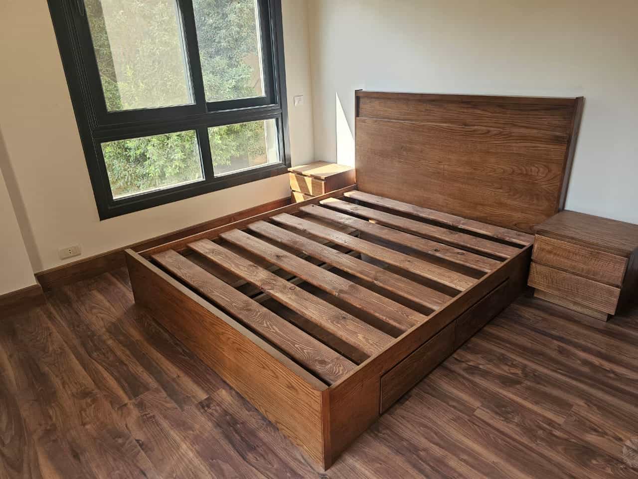Modern wooden bed frame in a bright, airy bedroom with large window overlooking greenery.